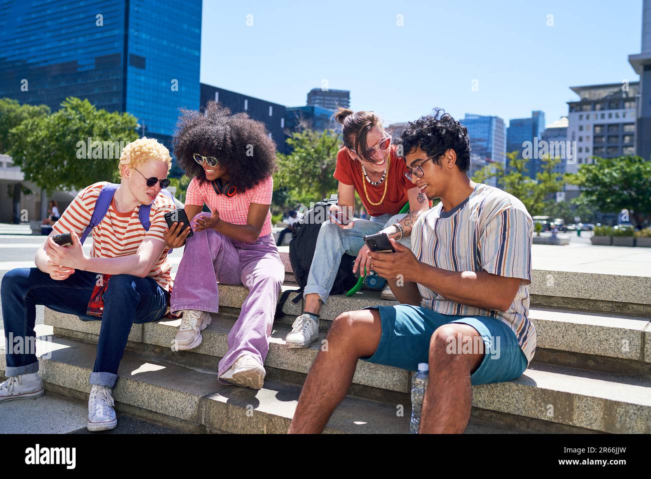 Giovani amici che si aggrappano, utilizzando gli smartphone nel soleggiato parco cittadino Foto Stock