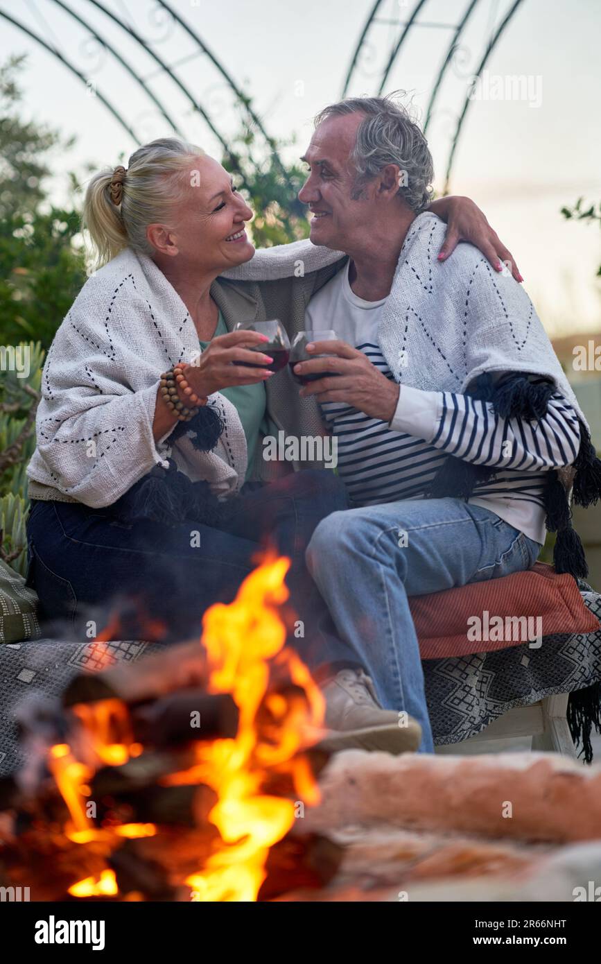 Felice coppia anziana che abbraccia e beve vino rosso dalla buca del fuoco sul patio Foto Stock