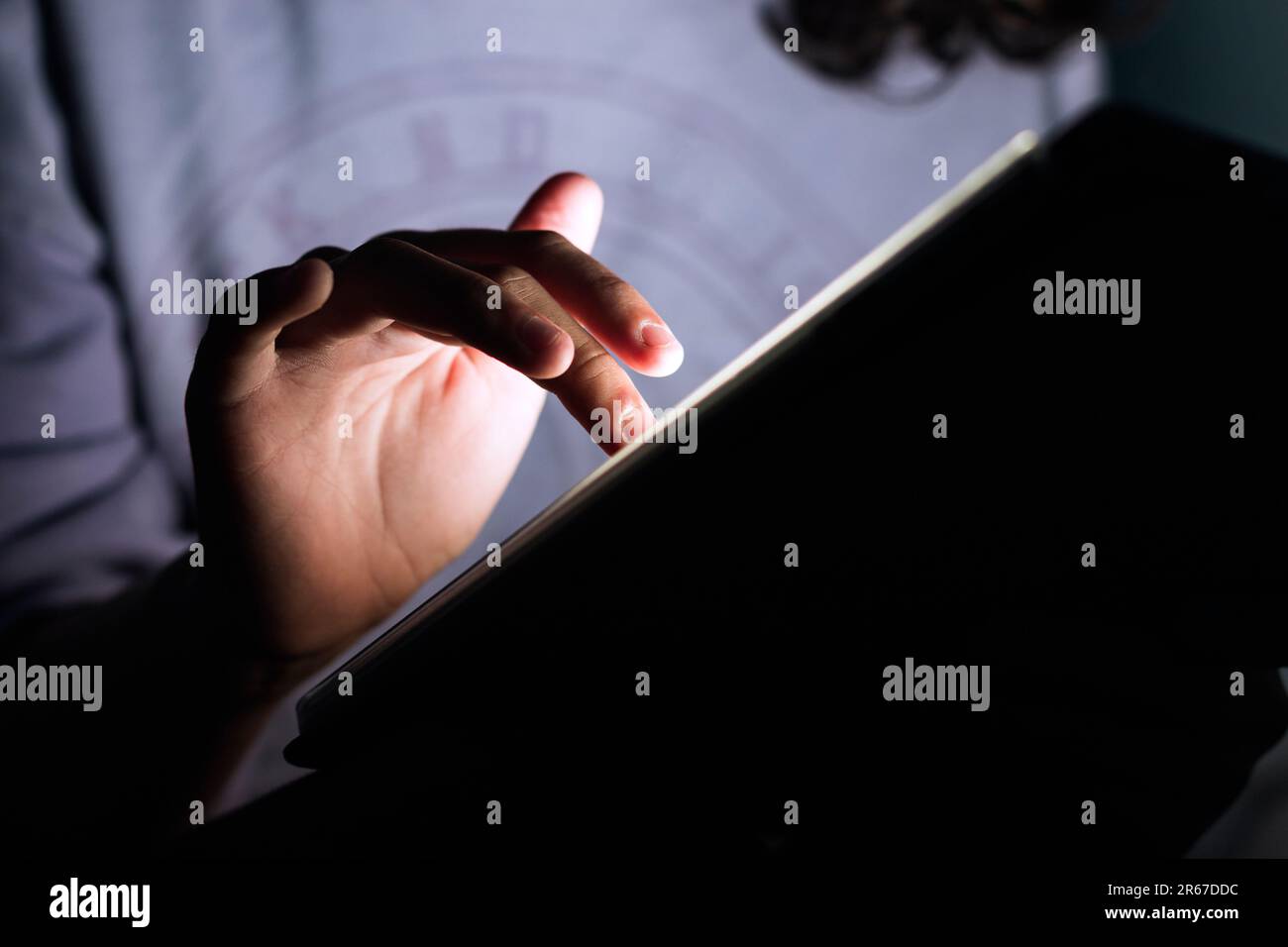 Primo piano di una mano di ragazza adolescente irriconoscibile che tocca lo schermo di un tablet digitale in una stanza buia. Foto Stock