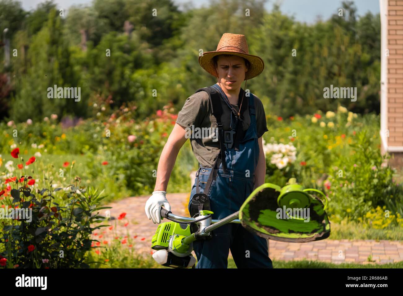 Un giovane uomo bianco in un cappello di paglia sta falciando un prato con un rasaerba nel suo giardino sul retro Foto Stock