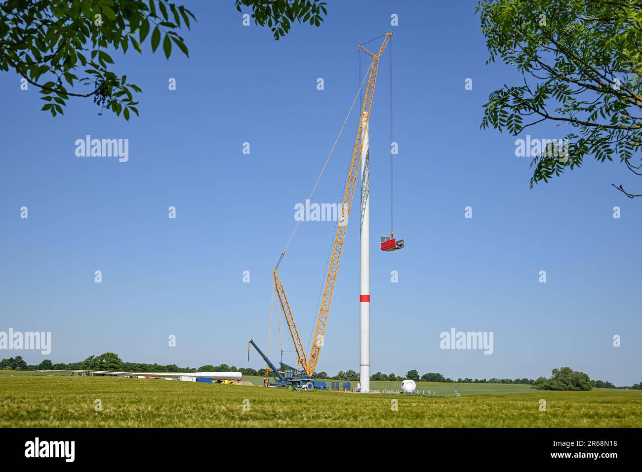 Installazione di una turbina eolica, gru alta che solleva la navicella sulla torre, mozzo del rotore, pale e generatore sdraiati sul cantiere, rinnovo Foto Stock