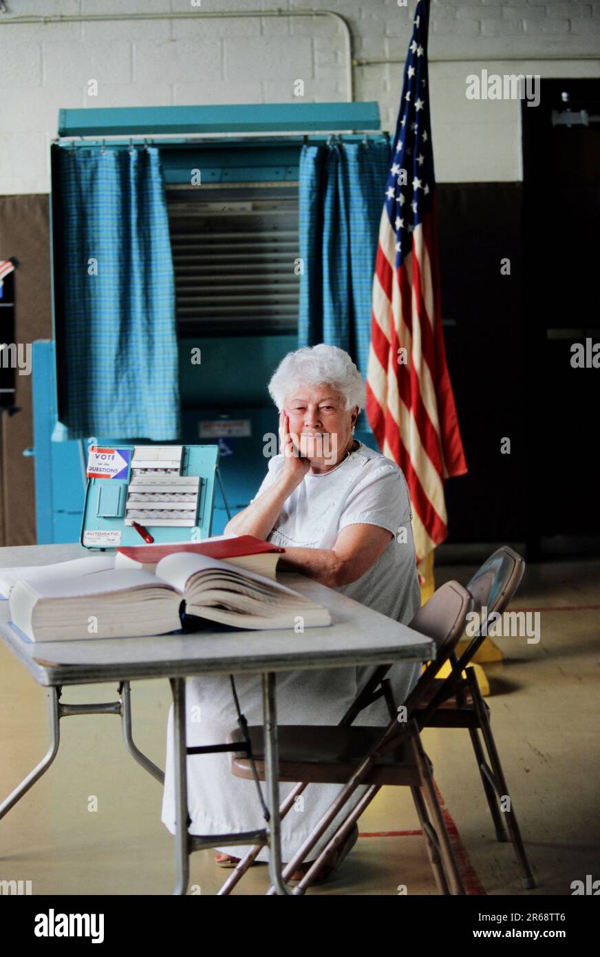 Ritratto di una donna anziana che si offre come funzionario elettorale in un luogo di scrutinio locale con bandiera americana e cabina di voto sullo sfondo Foto Stock