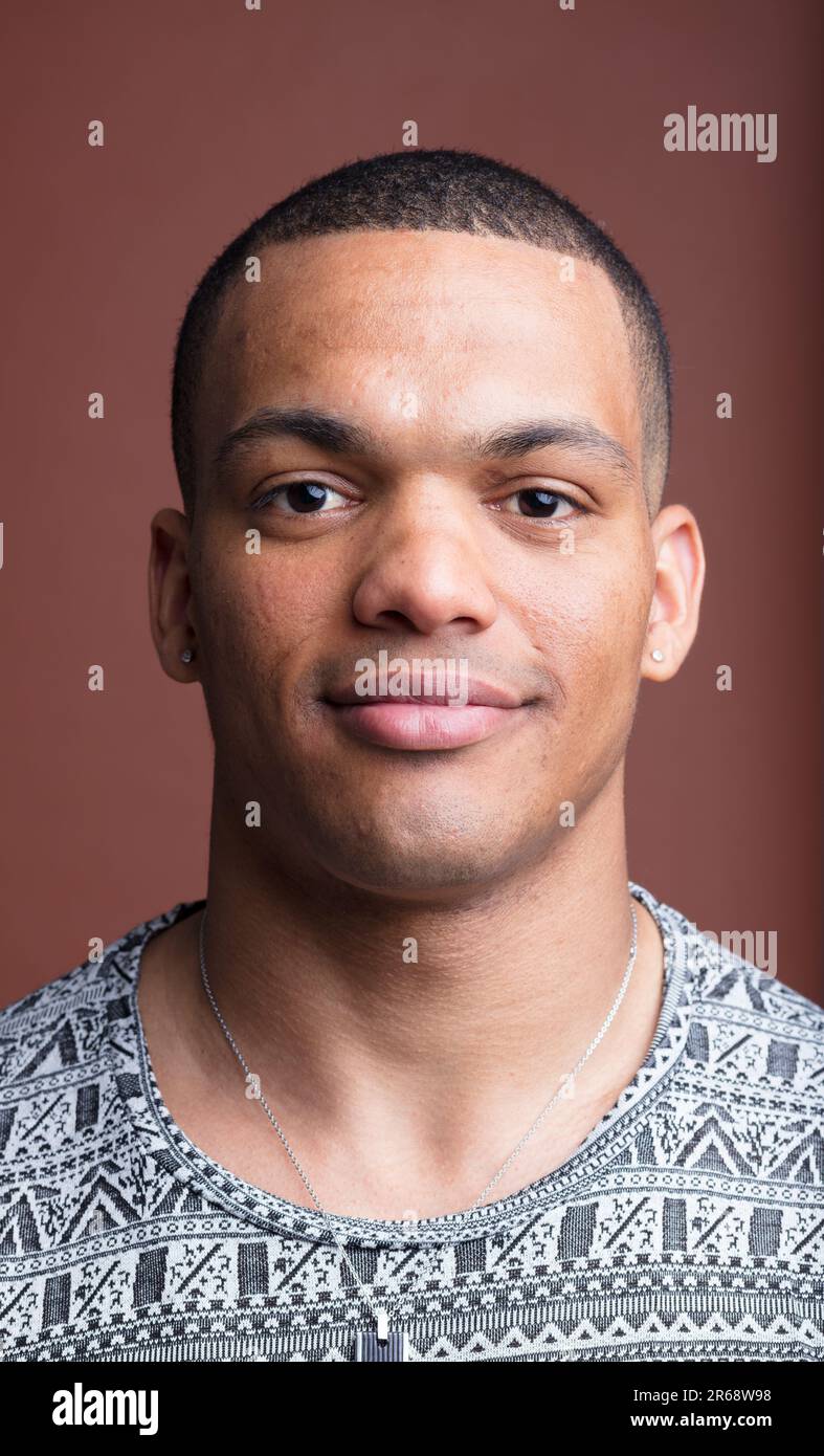 Giovane e robusto individuo afro-americano, sorridente, con testa rasata e piccolo orecchino a rombo, su sfondo marrone Foto Stock