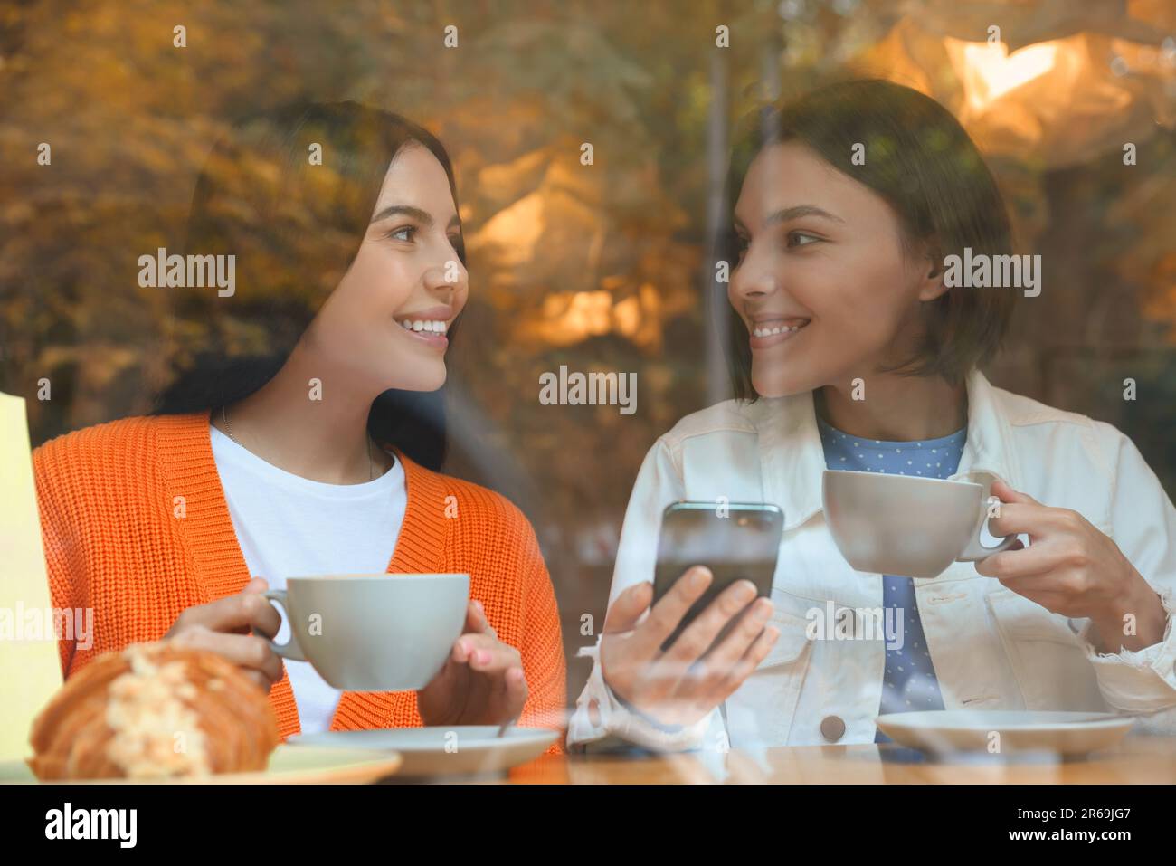 Promozione speciale. Donne giovani felici con tazze di bevande utilizzando smartphone al tavolo in caffè, vista dall'esterno Foto Stock Promozione speciale. Donne giovani felici con tazze di bevande utilizzando smartphone al tavolo in caffè, vista dall'esterno Foto Stock