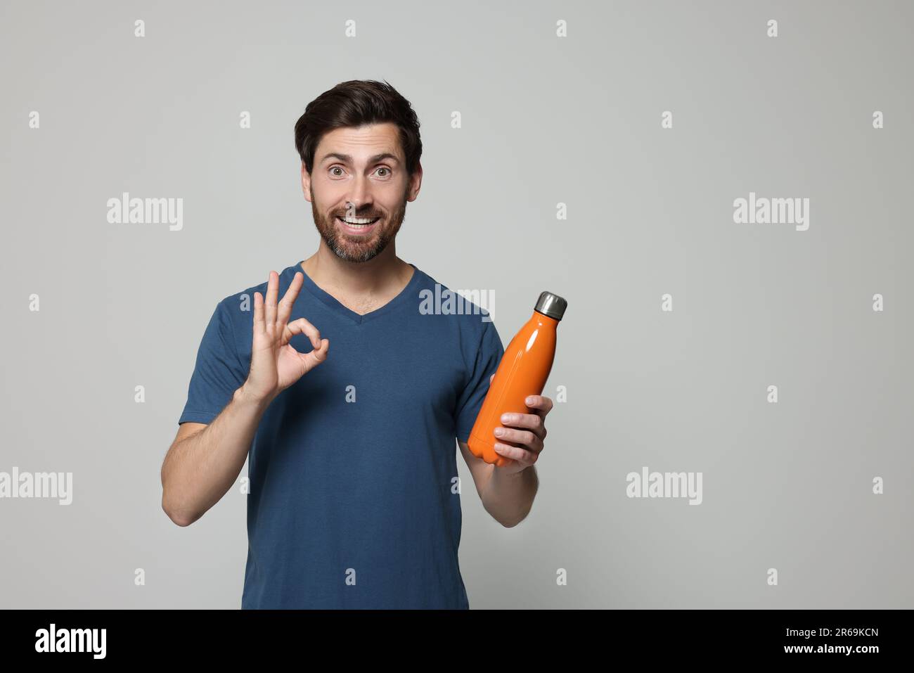 Uomo con bottiglia termica arancione con gesto ok su sfondo grigio chiaro. Spazio per il testo Foto Stock