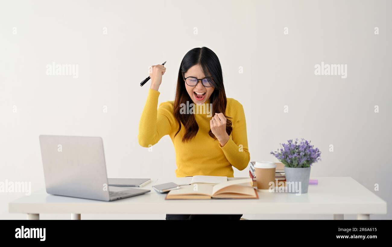 Una giovane studentessa asiatica di college, entusiasta e allegra, celebra un incredibile punteggio di esami e alza le braccia con pugni aggrappati a h Foto Stock