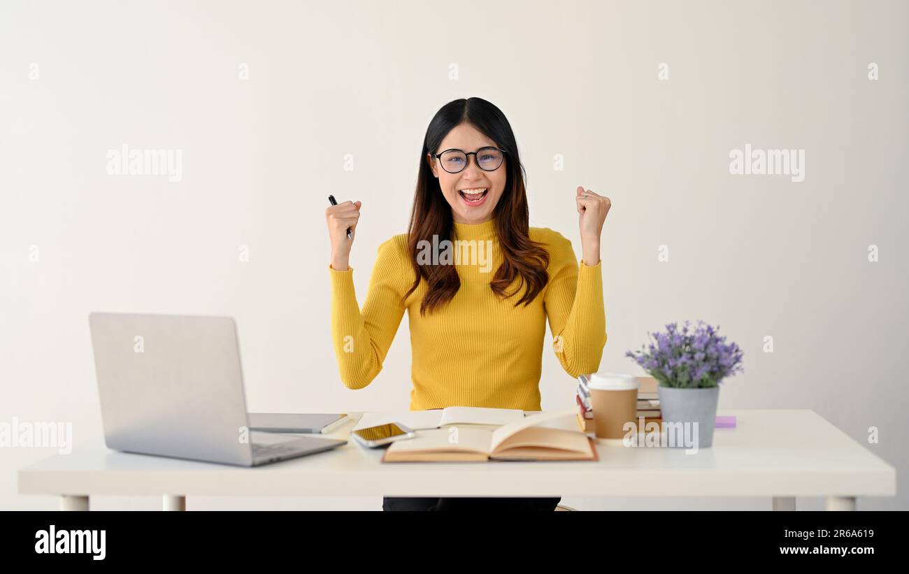 Una giovane ragazza asiatica allegra studentessa universitaria sta celebrando il suo successo e solleva le braccia con pugni aggrappati alla sua scrivania. isolato su una parte posteriore bianca Foto Stock