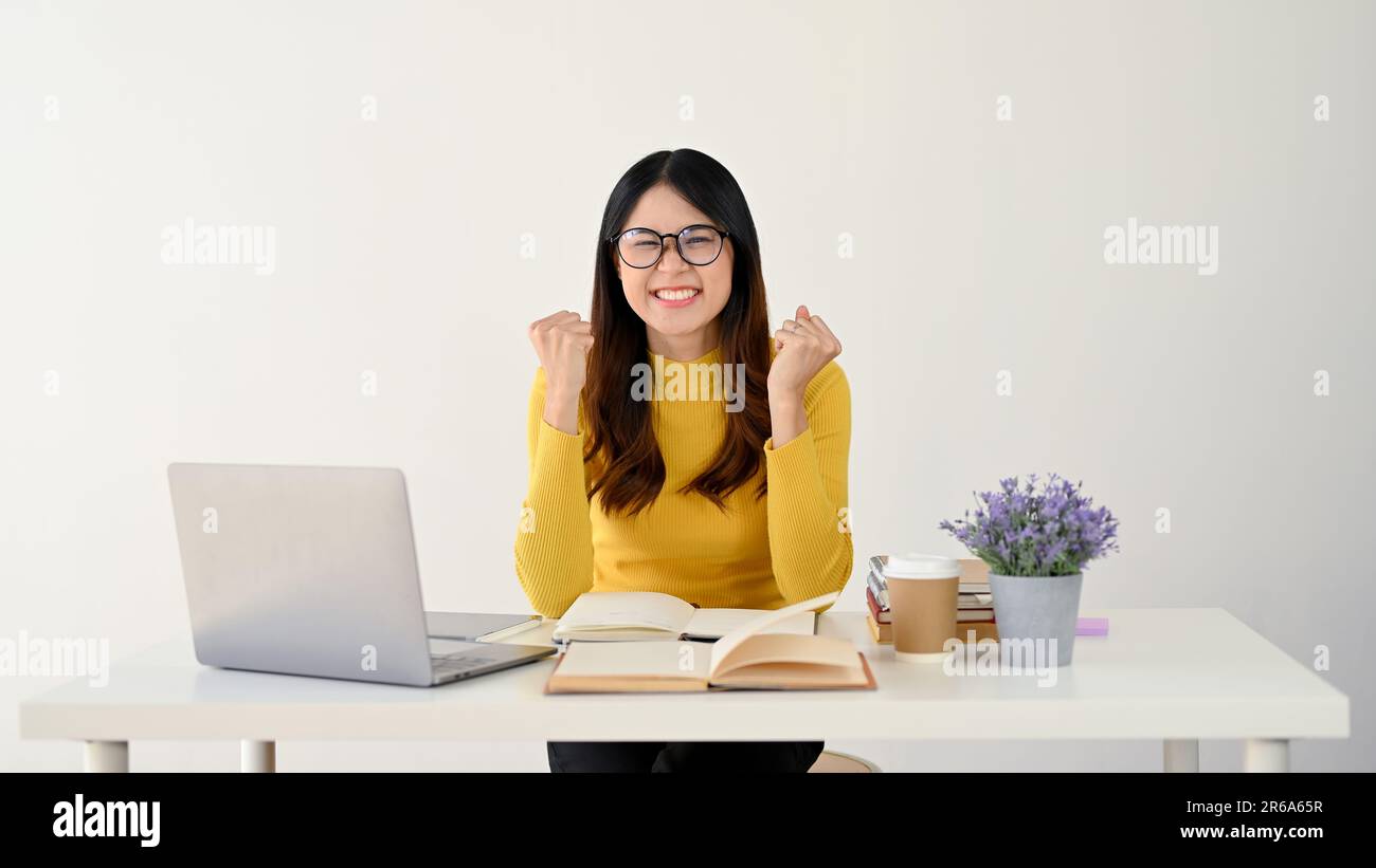 Una giovane studentessa asiatica di college entusiasta e allegra sta mostrando i pugni, gioendo e celebrando il suo punteggio finale mentre si siede a lei Foto Stock