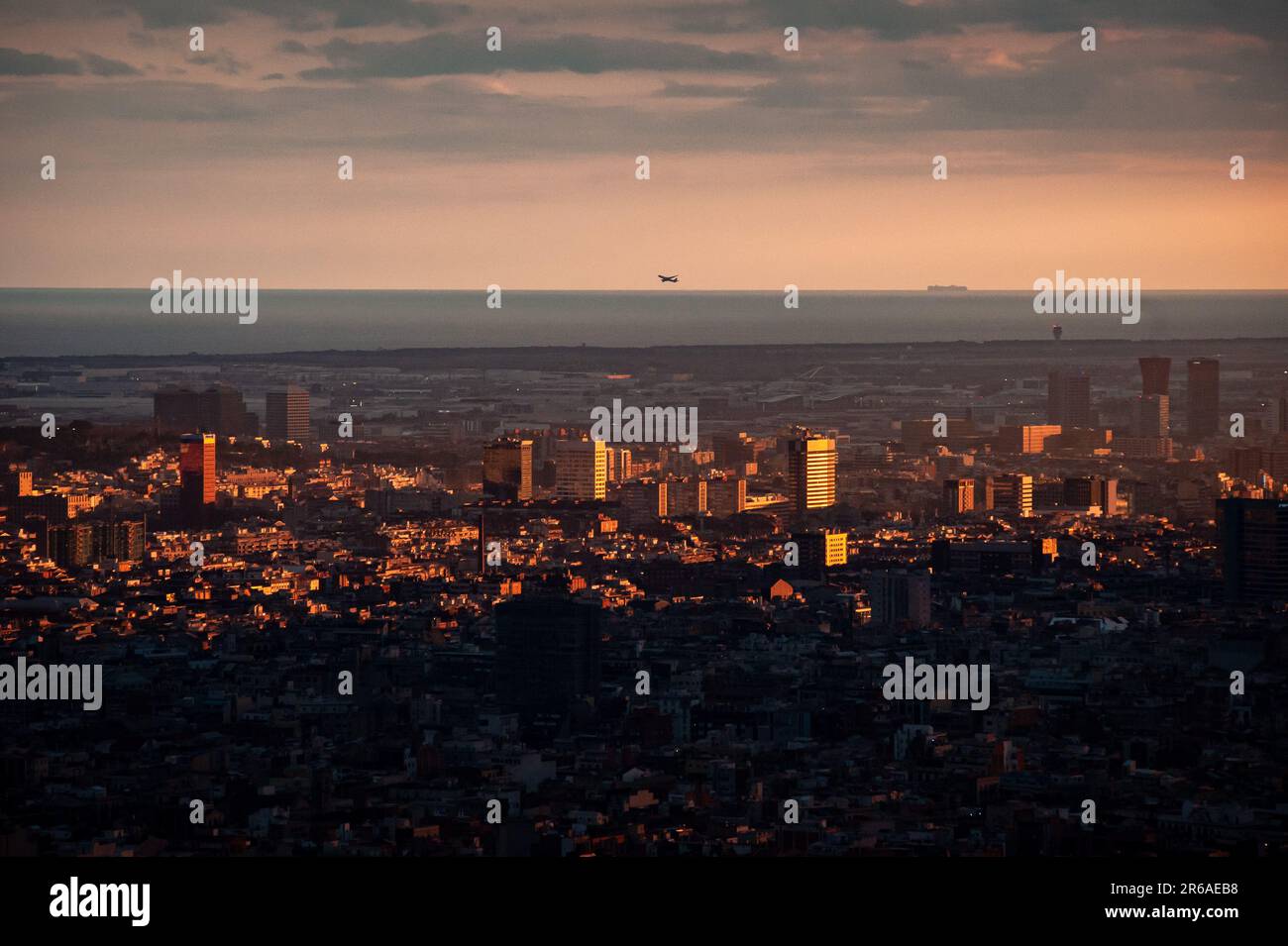 Vista aerea di Barcellona al tramonto, con lo skyline urbano illuminato dalla luce dorata, il Mar Mediterraneo all'orizzonte e un aereo in volo. Foto Stock