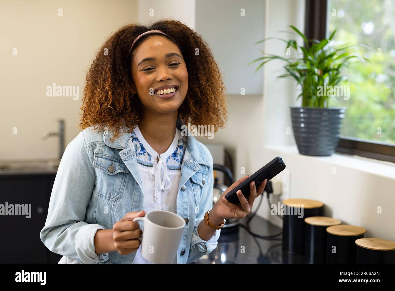 Ritratto di felice donna biraciale che ha il caffè, sorridendo e utilizzando smartphone in cucina a casa Foto Stock