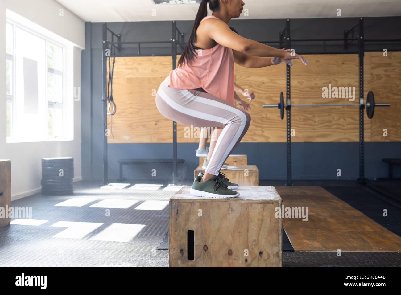 Vista laterale della giovane donna biraciale che pratica squat su plyo box in legno in palestra, spazio copia Foto Stock