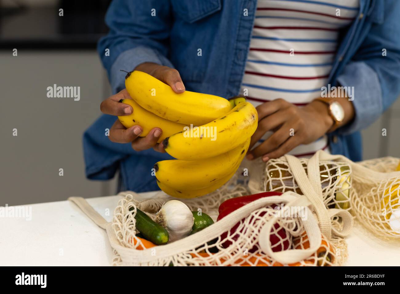 A metà della donna biraciale disimballare i sacchetti di shopping della drogheria in cucina che tiene le banane Foto Stock