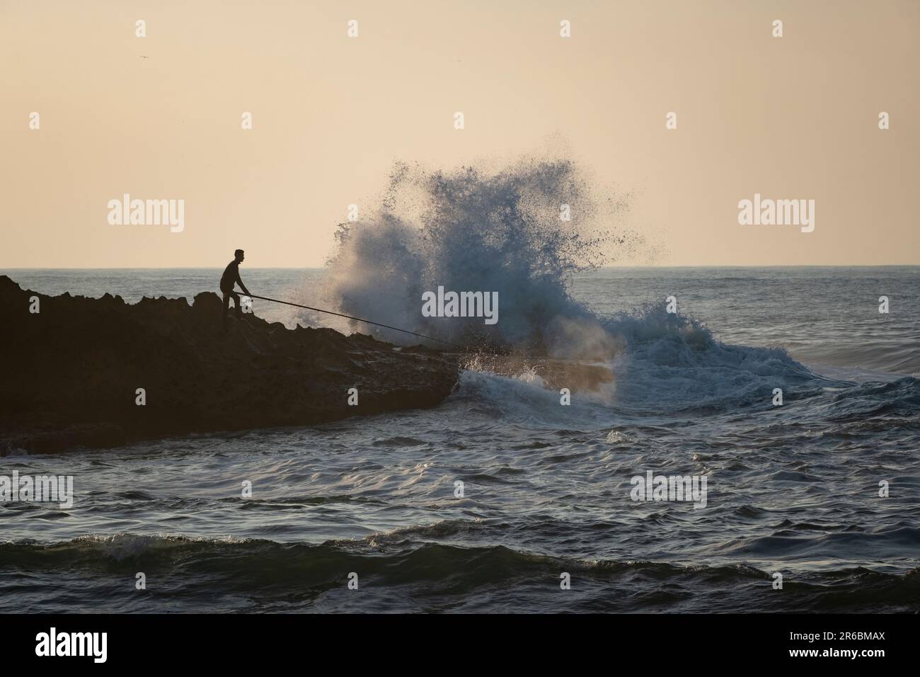 Un pescatore silhouette che si erge su uno sperone roccioso dell'oceano Atlantico con la sua canna da pesca rivolta verso le onde che si infrangono a Oualidia, Marocco Foto Stock