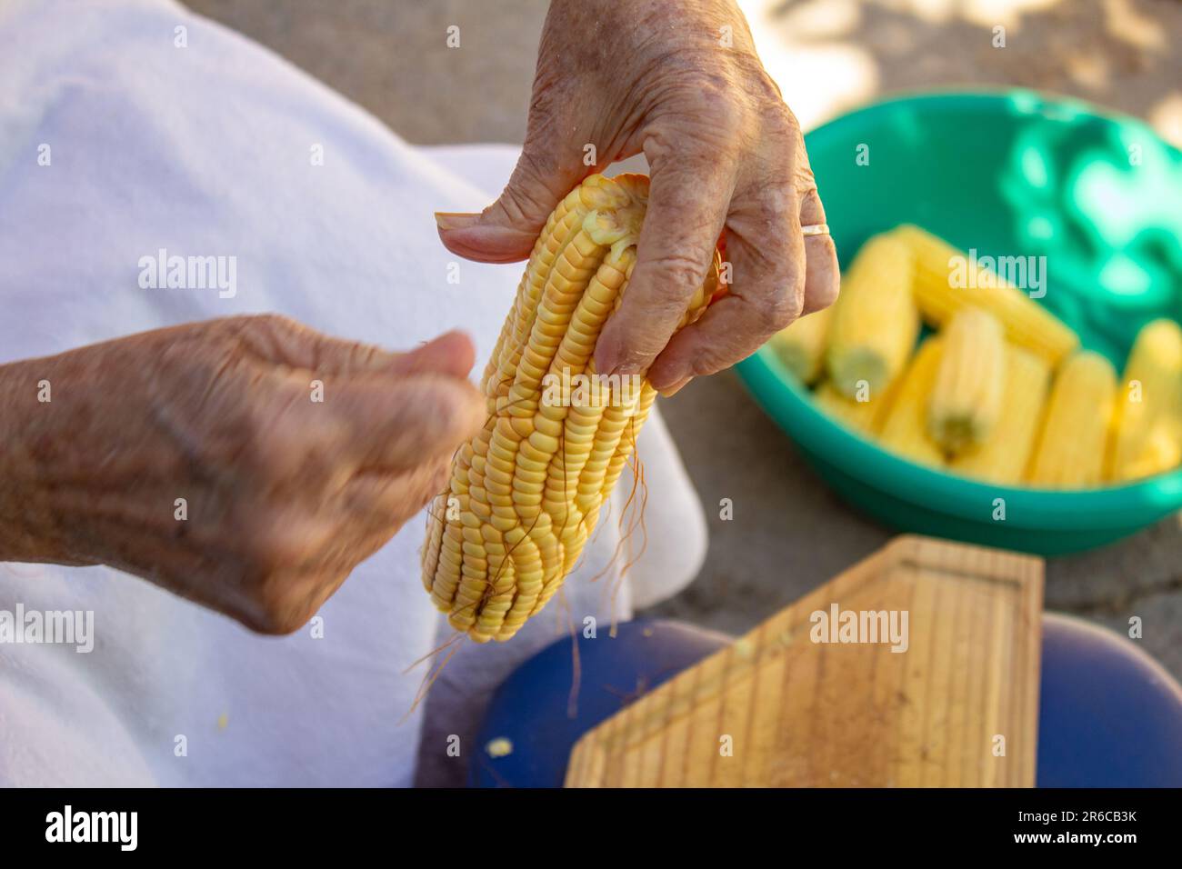 Goiania, Goias, Brasile – 02 giugno 2023: Le mani di una donna che tira i capelli dal mais verde e una ciotola verde piena di pannocchie di mais sullo sfondo. Foto Stock