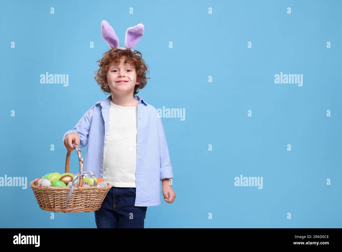 Ragazzo felice in cute coniglietto orecchie fascia che tiene il cestino di vimini con uova di Pasqua su sfondo azzurro. Spazio per il testo Foto Stock