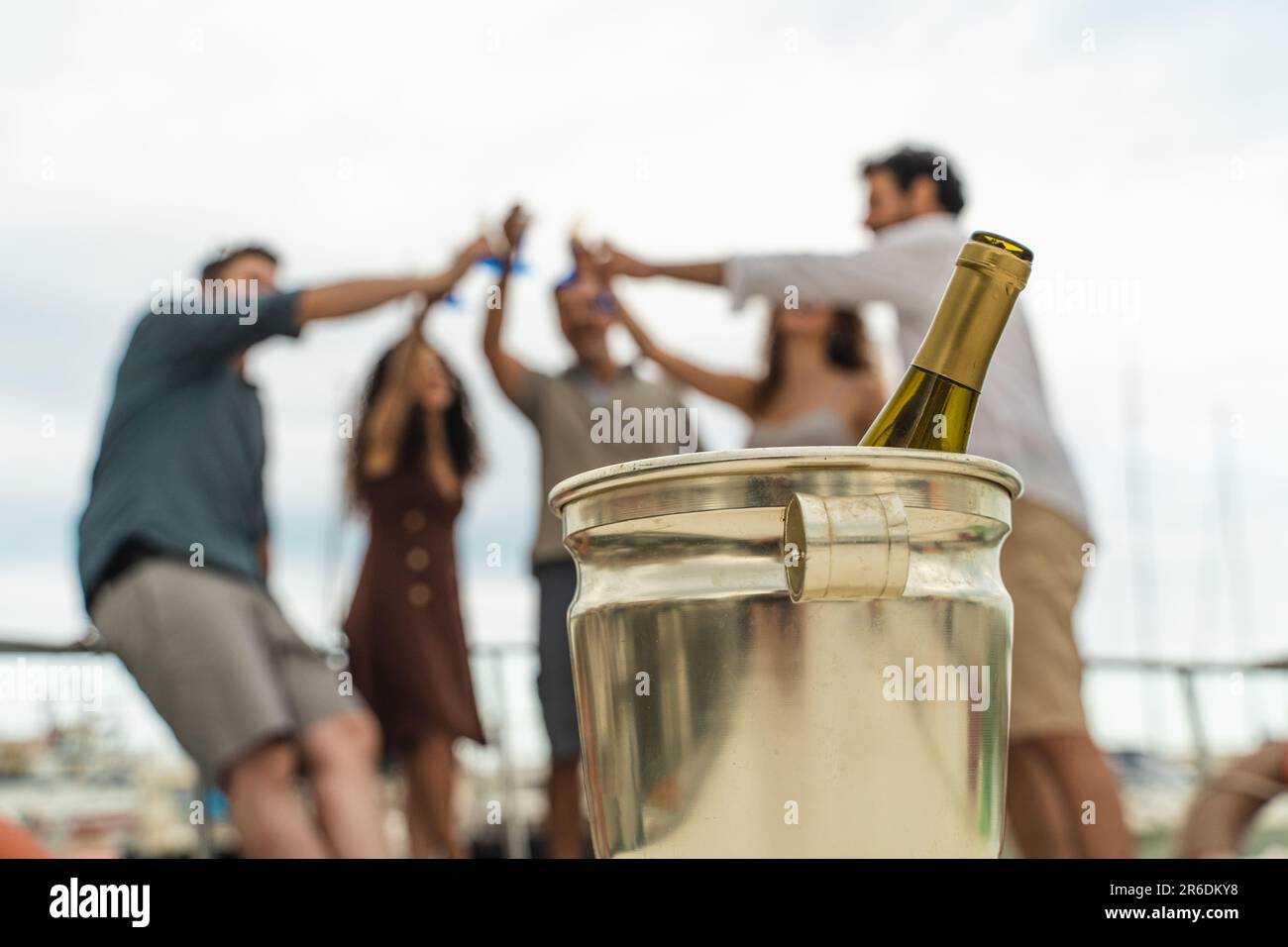 Primo piano di una bottiglia di vino frizzante in un secchio di ghiaccio mentre un gruppo di amici brinda allegramente sullo sfondo. Gli amici, fuori fuoco, c Foto Stock