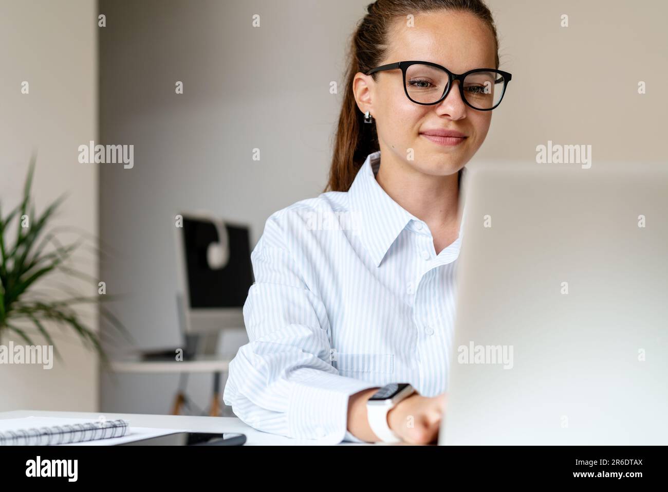 Giovane bruna donna d'affari in occhiali sorridente e al lavoro su un computer portatile in ufficio Foto Stock