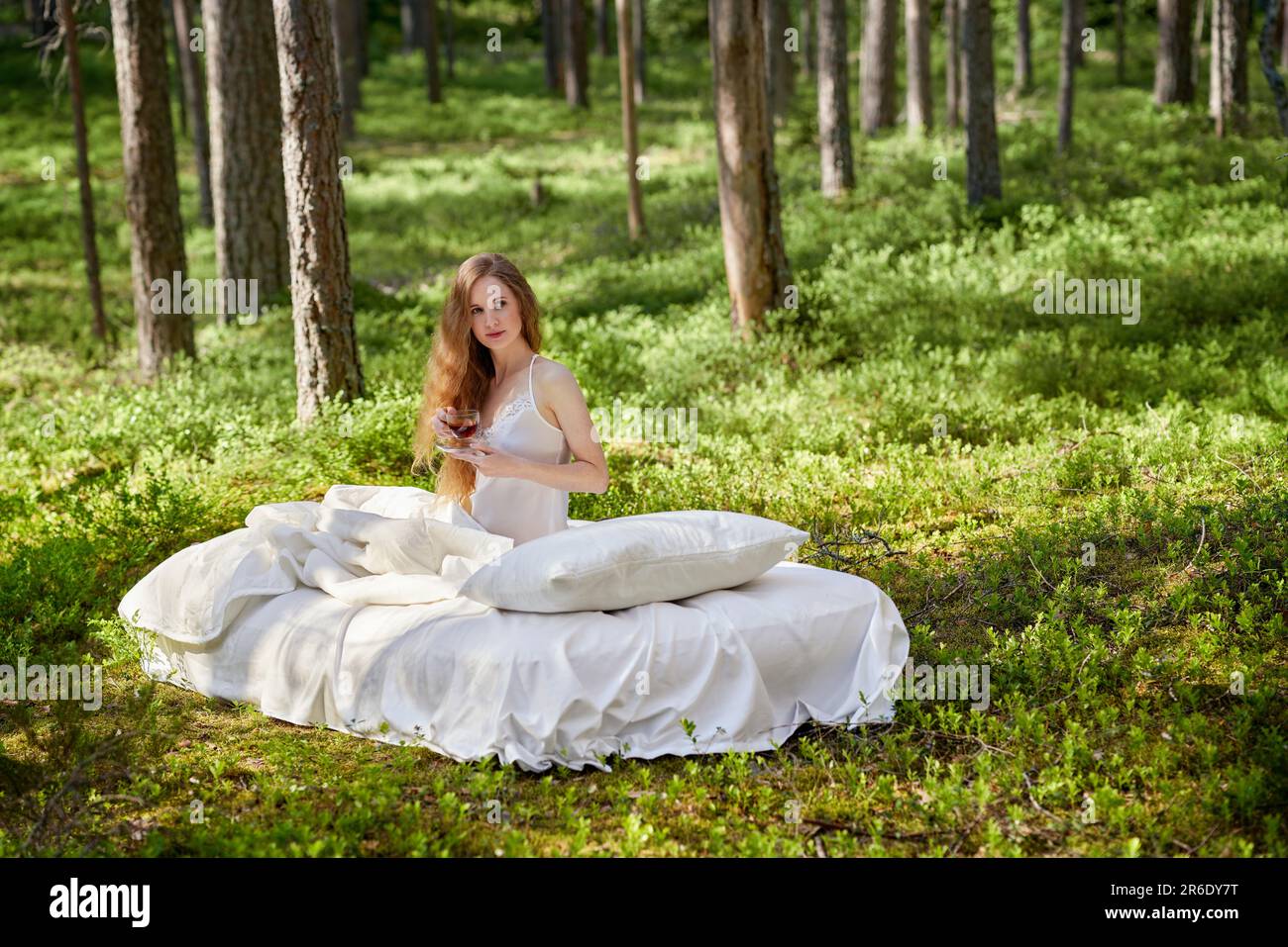 La donna si sveglia in una foresta estiva. Una ragazza beve il tè del mattino seduto su un materasso nel bosco Foto Stock