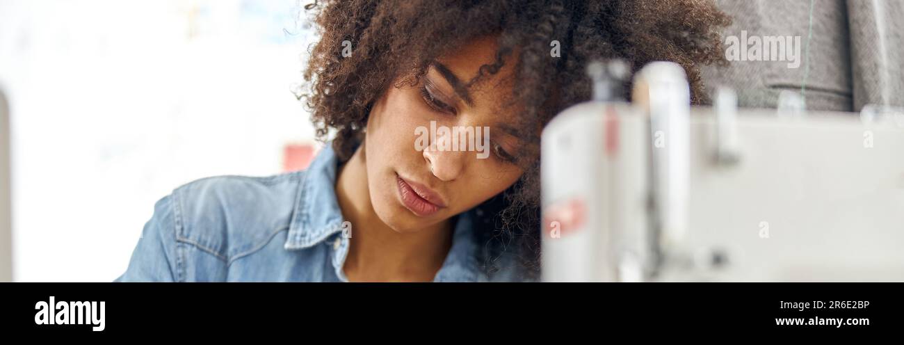 Il dressaker afroamericano concentrato si ispira a un libro di schizzo seduto sul posto di lavoro con una moderna macchina da cucire Foto Stock