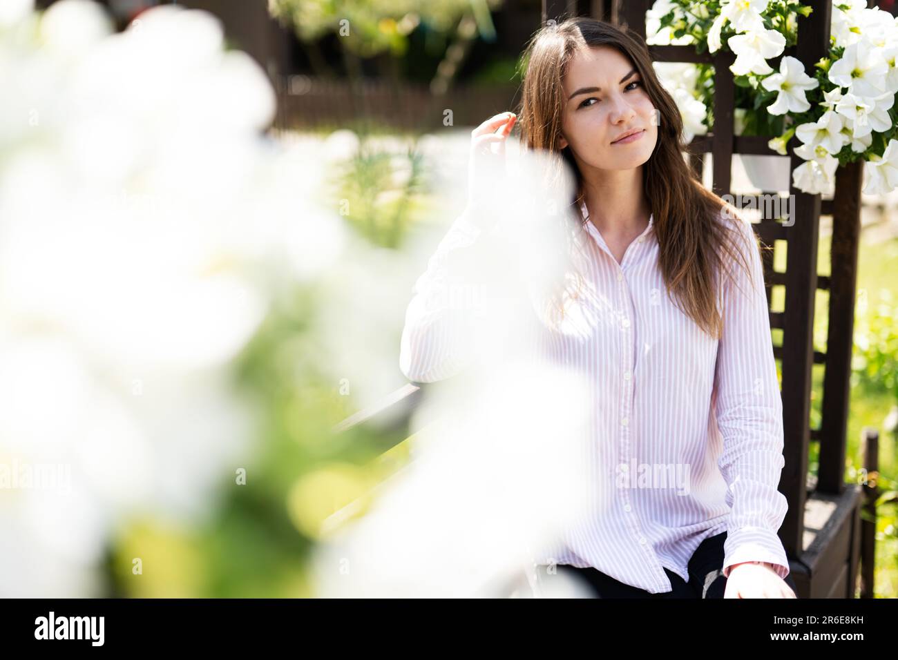 Giovane bella donna in camicia seduta su panca di legno con fiori in giardino. Foto Stock