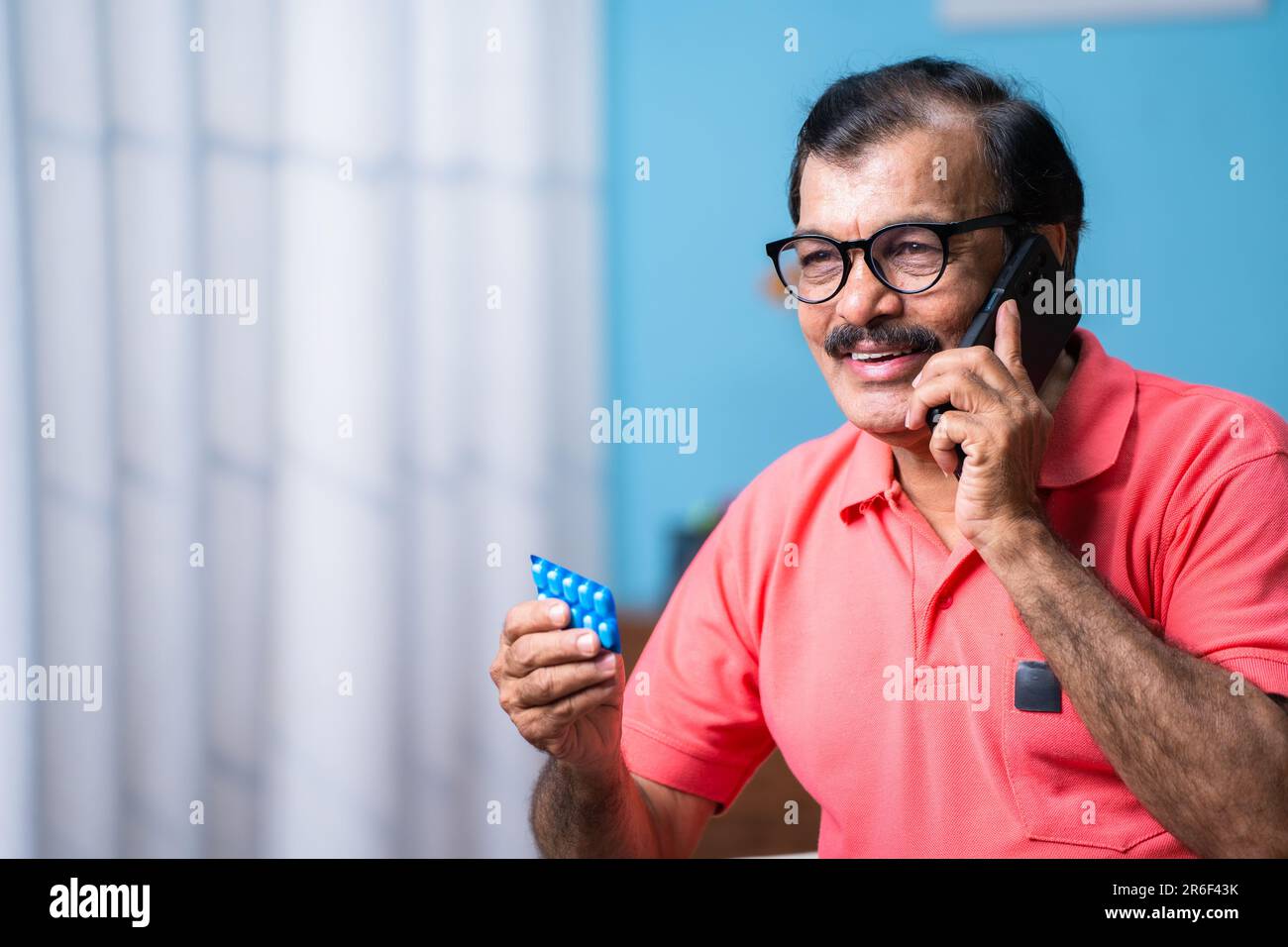 Uomo anziano indiano che parla sul telefono cellulare tenendo i tablet a portata di mano a casa - concetto di telemedicina, assistenza sanitaria anziana e farmacia Foto Stock