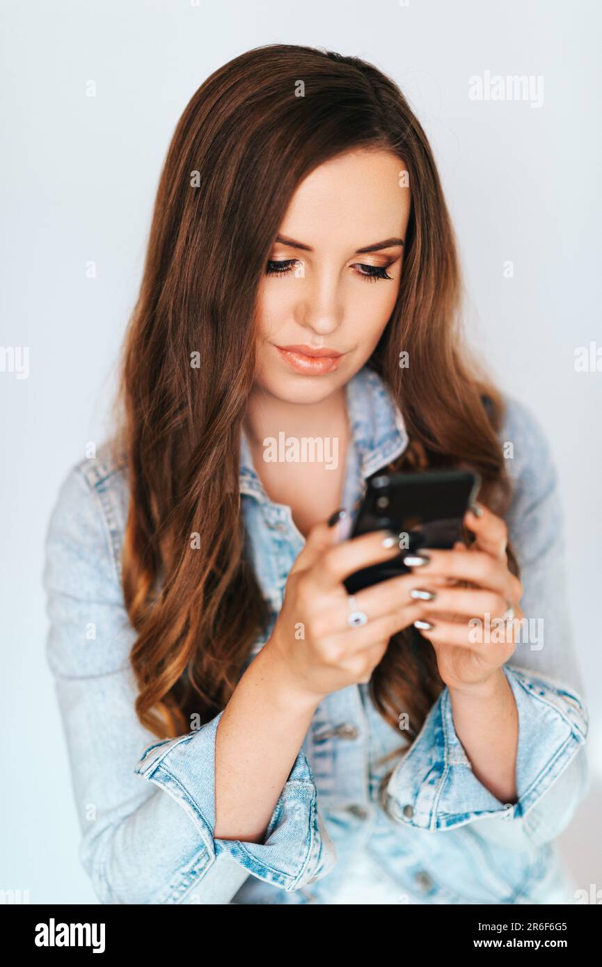 Ritratto di giovane bella donna con lunga acconciatura, indossare giacca blu denim, messaggio di testo sul telefono Foto Stock