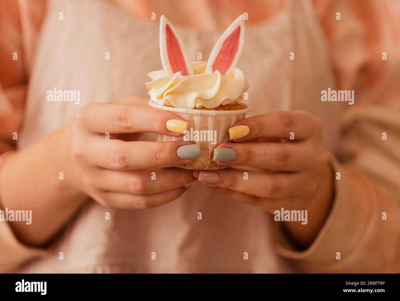 Primo piano delle mani femminili con una manicure ordinata tenere una torta per bambini. Delizioso cupcake carino con orecchie di coniglio e crema cagliata. Abiti donna di colore beige su t Foto Stock