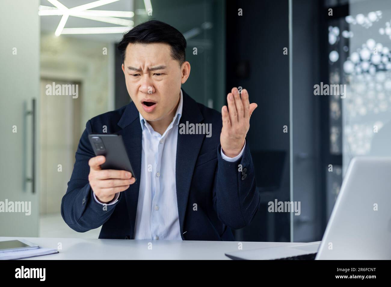 Un ragazzo asiatico confuso che tiene in mano uno smartphone si sente preoccupato di pensare al messaggio ricevuto. Utilizzo quotidiano del telefono cellulare, notizie spiacevoli, attesa di una chiamata importante, concetto di problemi di dispositivo con segnale basso Foto Stock