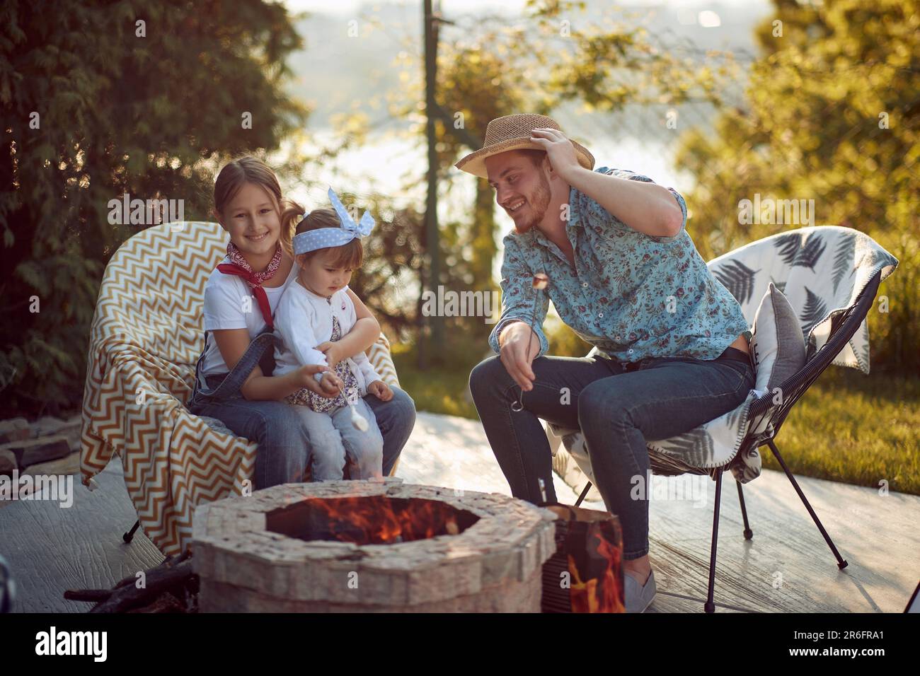 Gioiosa giovane ragazze arrostire marshmallows da un camino con il loro papà all'aperto in un cottage accogliente, godendo di sole giornata estiva insieme. Stile di vita, Toge Foto Stock