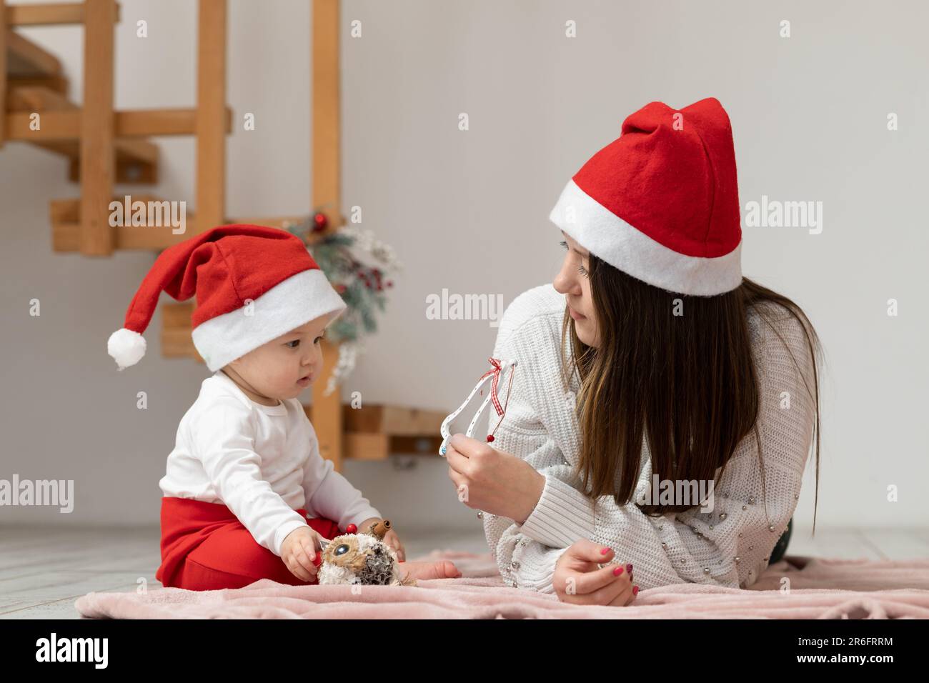 Una giovane donna gioca con sua figlia sdraiata su una coperta disposta sul pavimento e le offre di prendere un giocattolo di Capodanno. Foto Stock