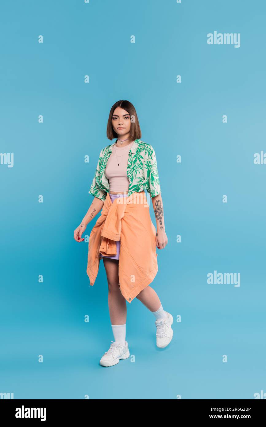 stile di tutti i giorni, giovane donna brunetta con capelli corti che cammina in camicia con stampa di palme, gonna e sneaker bianche su sfondo blu, spensierato, tat Foto Stock