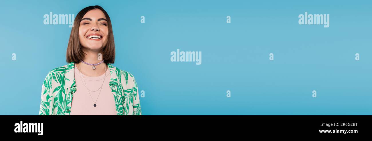 giovane donna allegra con capelli corti brunette che indossa una camicia con stampa palma, sorridente con occhi chiusi su sfondo blu, abbigliamento casual, gen z fa Foto Stock