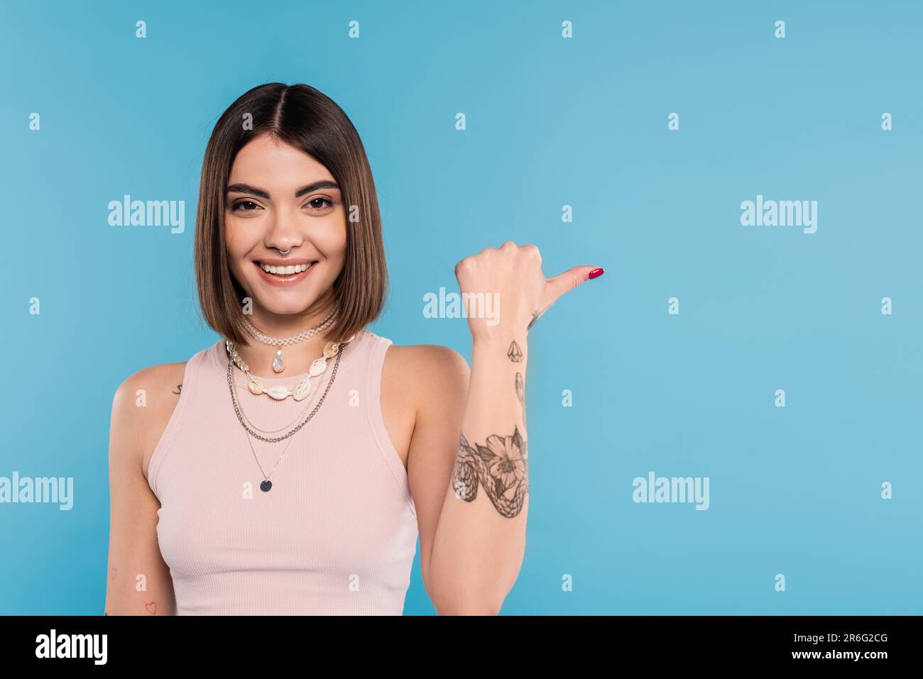 allegria, tatuata giovane donna con capelli corti brunette in canotta top sorridente e puntando con pollice su sfondo blu, abbigliamento casual, gen z fishi Foto Stock