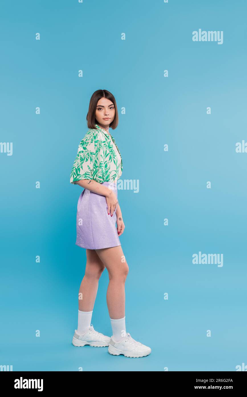 moda estiva, giovane donna brunetta con capelli corti in camicia con stampa palma, gonna e sneaker bianche su sfondo blu, spensierata, tatt Foto Stock