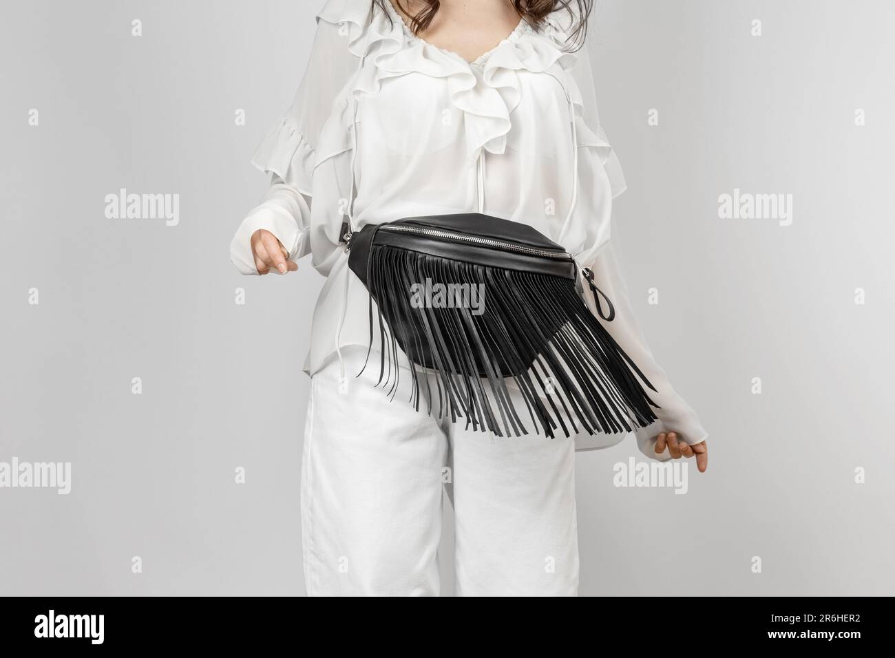 Donna bianca che indossa jeans bianchi, blusa con fronzoli e borsa a tracolla in pelle nera con frangia decorativa su sfondo grigio Foto Stock