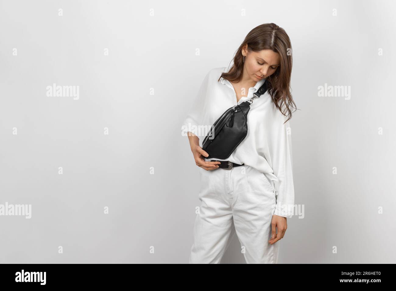 Donna bianca che indossa jeans bianchi, camicia e tracolla Borsa da cintura in pelle nera su sfondo grigio Foto Stock