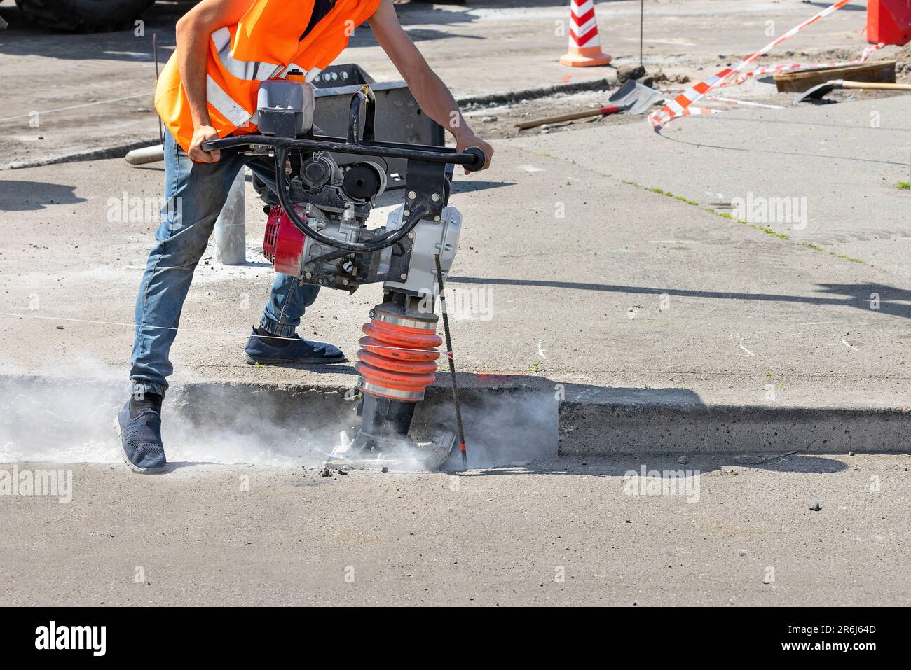 Un operaio di costruzioni sta facendo una trincea con un pestello vibrante in un cantiere edile e sta dando il via a una nuvola di polvere intorno a lui. Foto Stock
