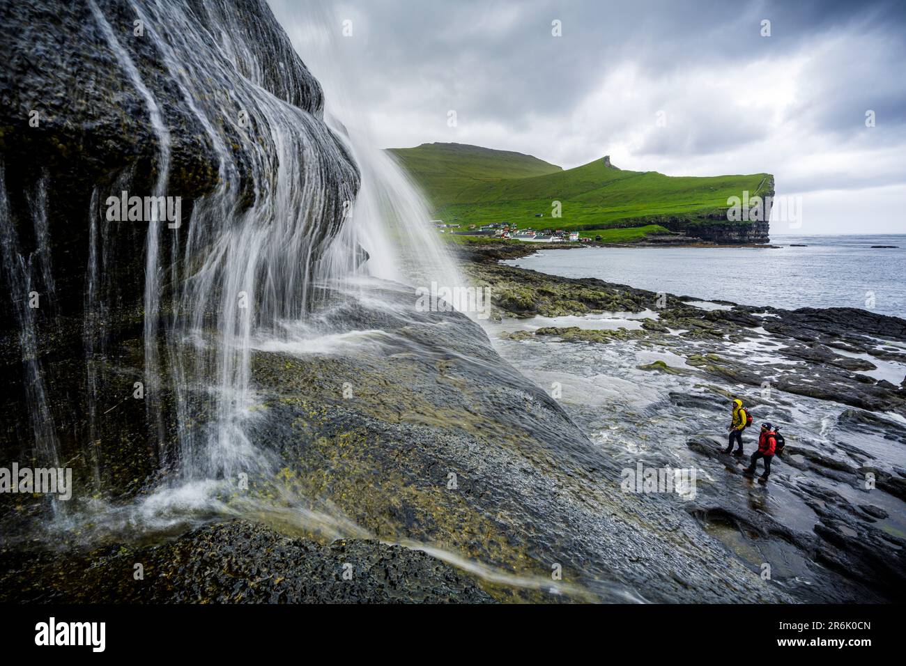 Vista ad angolo alto di due escursionisti che ammirano una maestosa cascata che si erge sulle scogliere, Gjogv, Isola di Eysturoy, Isole Faroe, Danimarca, Europa Foto Stock