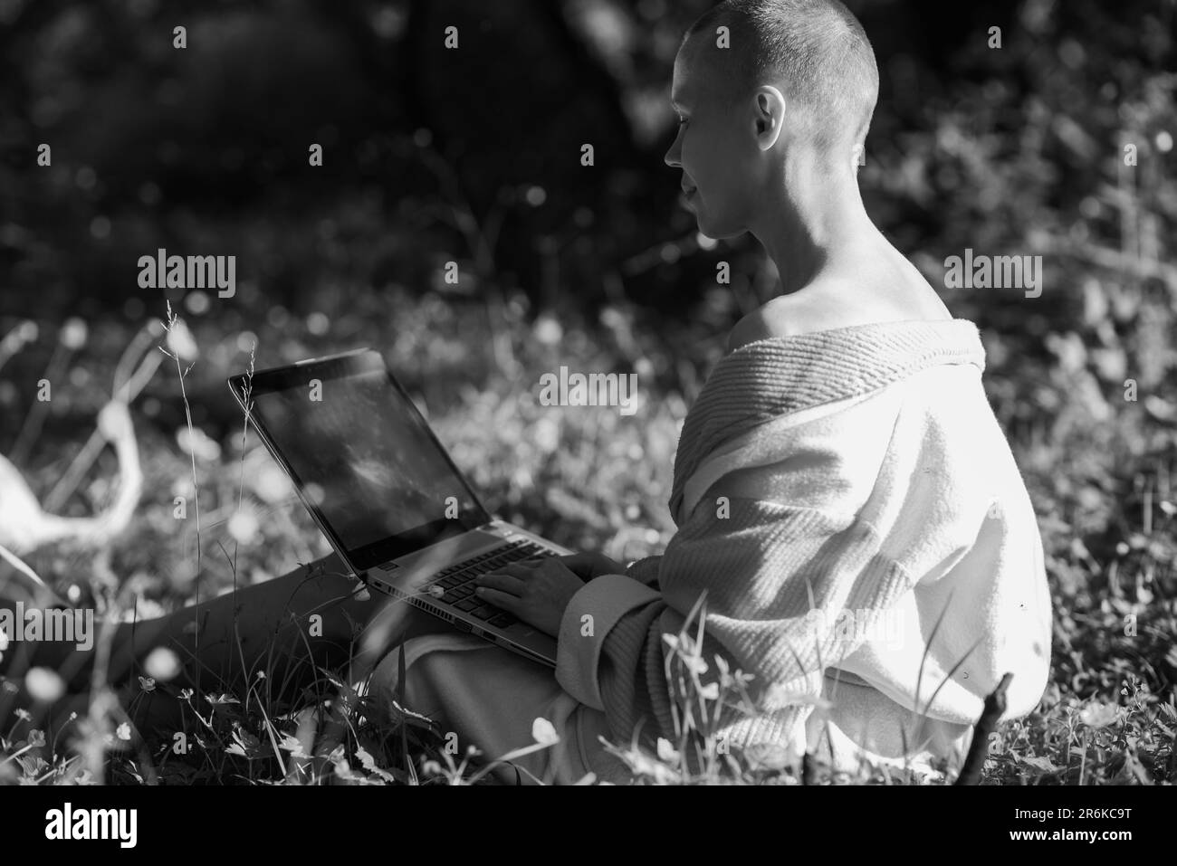Una donna calva in un accappatoio si siede sull'erba verde con un computer portatile in mano. Lavoro online, concetto di freelance. Giorno di sole. Immagine in bianco e nero. Foto Stock