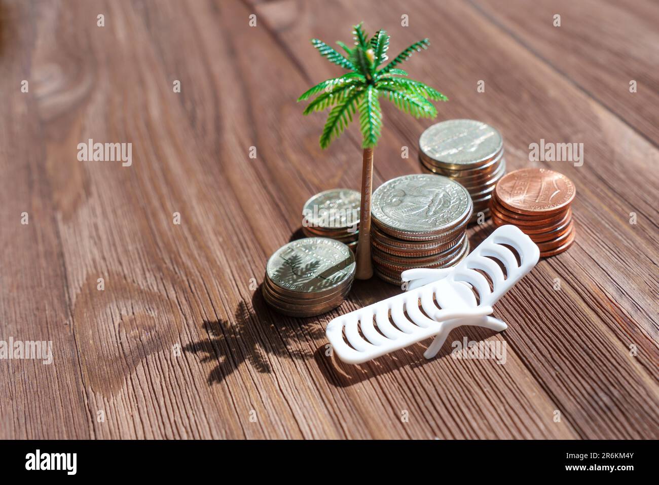 Nomadi digitali: Pile di monete americane, un lettino da spiaggia giocattolo e una palma disposti su uno sfondo di legno. Libertà e flessibilità di lavorare Foto Stock