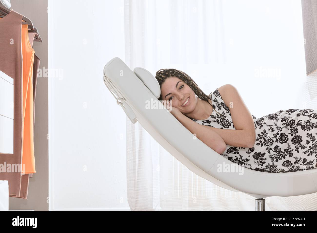 Una giovane donna rilassata riposa su una chaise lounge di design a forma di mezzaluna in una sala luminosa. Ha i capelli delle trecce della scatola e un vestito bianco e nero Foto Stock