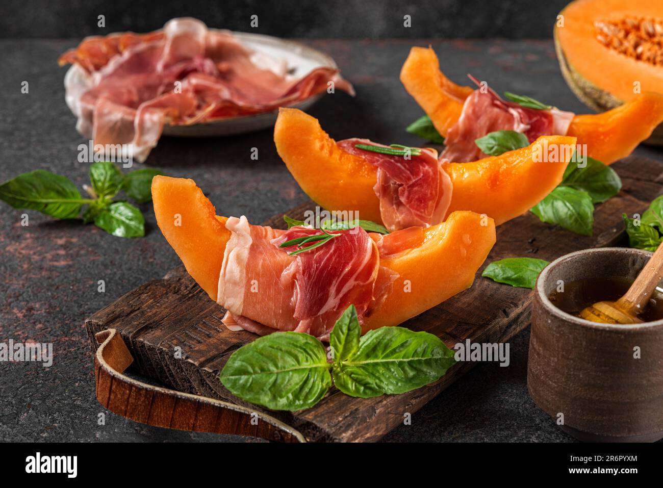 Prosciutto con fettine di melone cantaloupe, miele e basilico sul tagliere su fondo scuro. Antipasto italiano Foto Stock