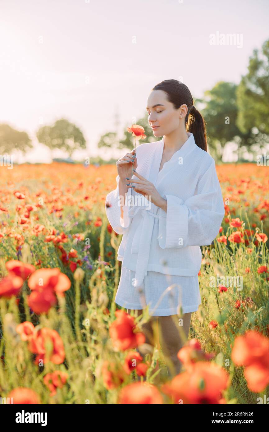 Giovane donna in abiti bianchi che cammina sul prato e che si gode da papaveri fioriti con gli occhi chiusi al tramonto. Foto Stock