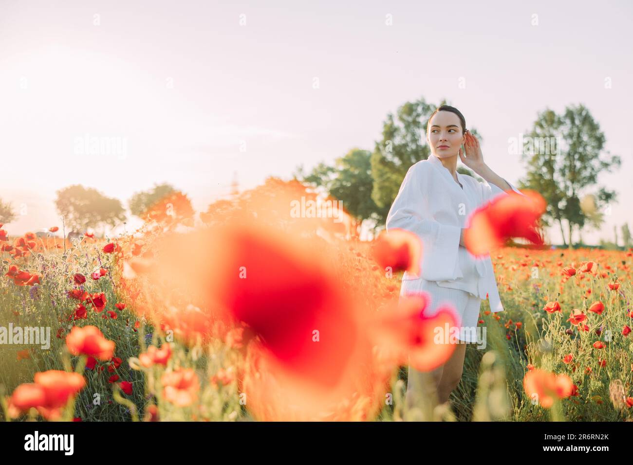 Giovane donna in abiti bianchi che cammina e si gode tra poppies fiorito prato al tramonto. Immagine con spazio di copia. Foto Stock