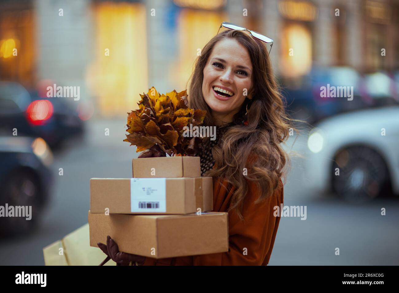 Ciao ottobre. felice donna di mezza età in trench arancione con pacchi e foglie gialle autunnali in città. Foto Stock