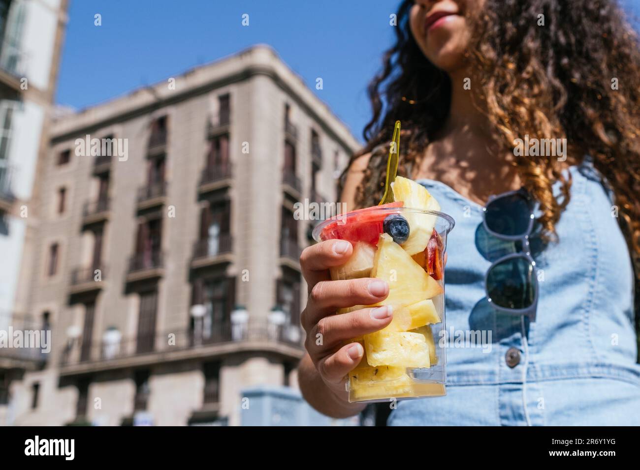 Morbido fuoco di anonima giovane donna che porta la tazza di plastica con pezzi di frutta fresca durante la passeggiata sulla strada della città illuminata dal sole Foto Stock