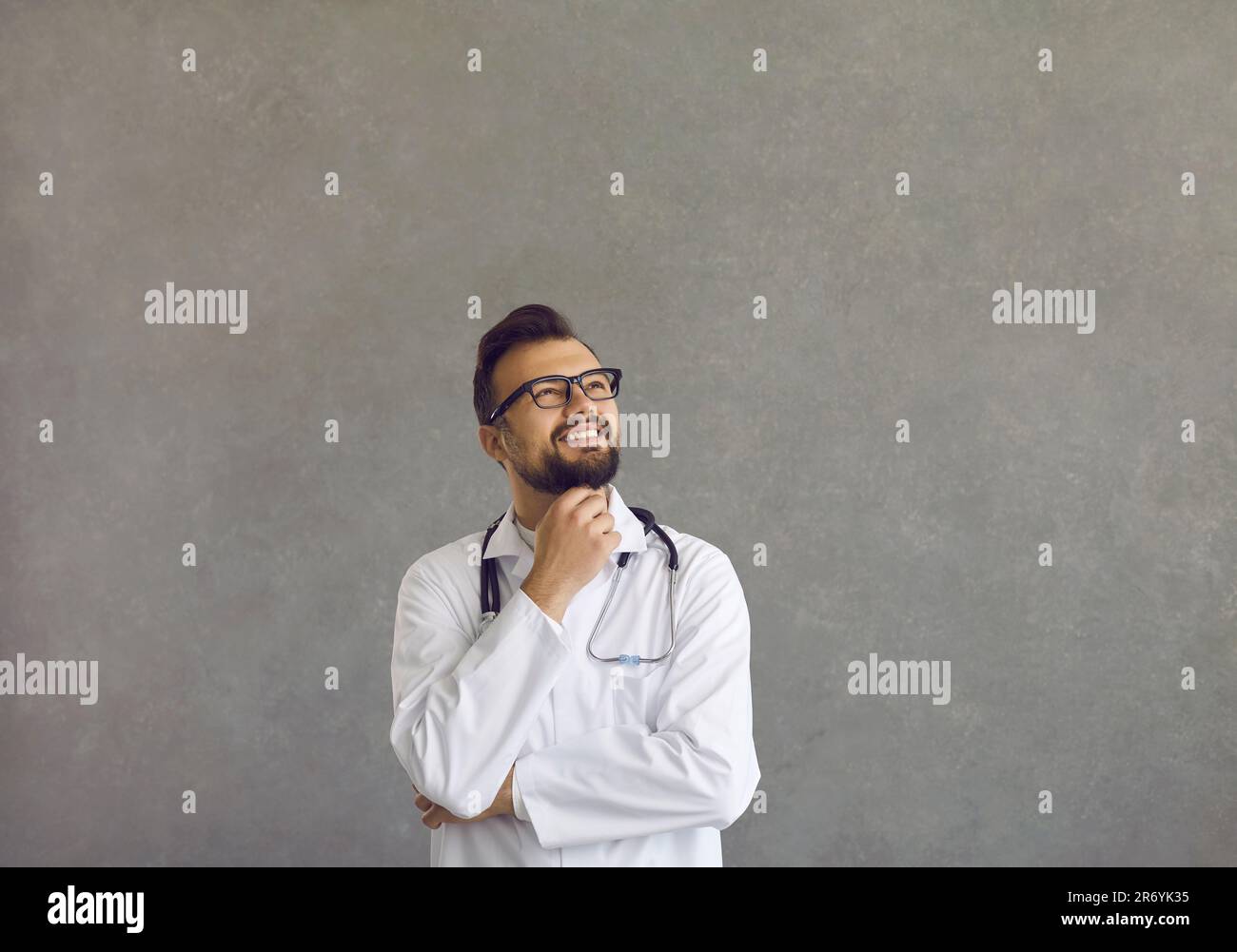 Soddisfatto medico maschio in cappotto bianco guardando su mento toccante sorridente studio scatto Foto Stock