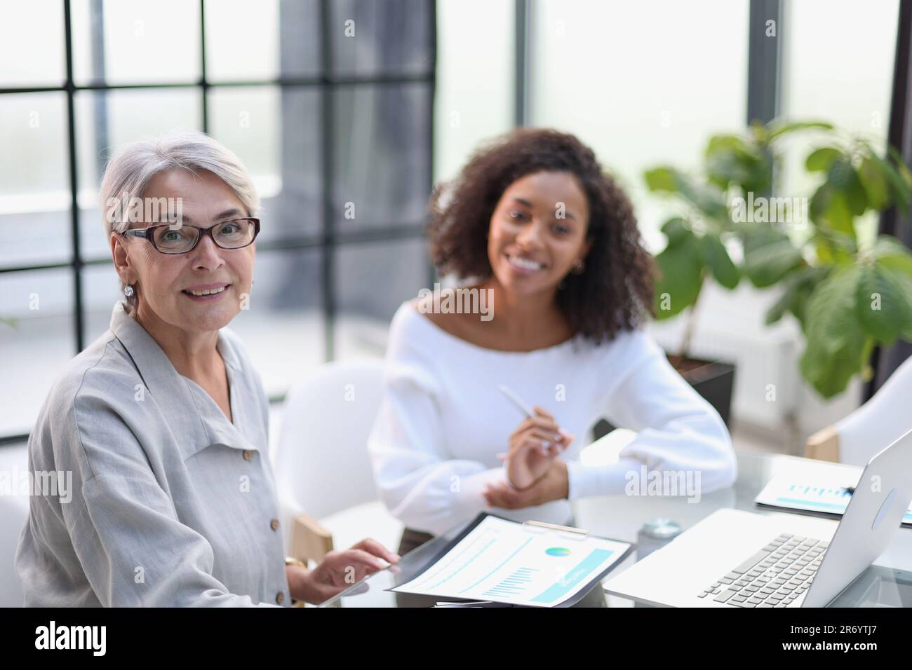 Due donne in viaggio sorridono mentre lavorano insieme su un computer portatile a un tavolo nella sala riunioni dell'ufficio Foto Stock
