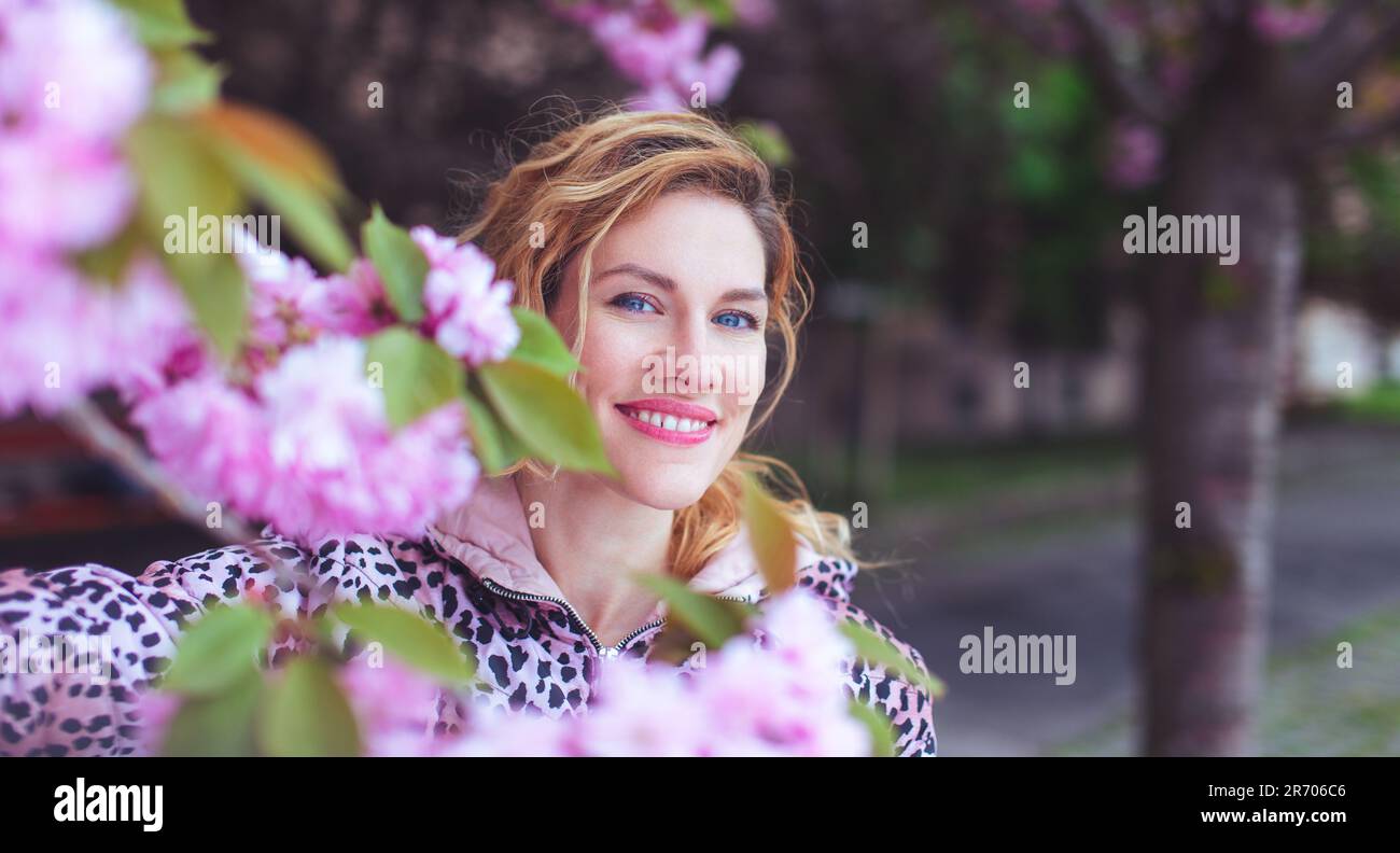 Felice 30s donna che guarda in macchina fotografica nel parco durante la fioritura del ciliegio Foto Stock