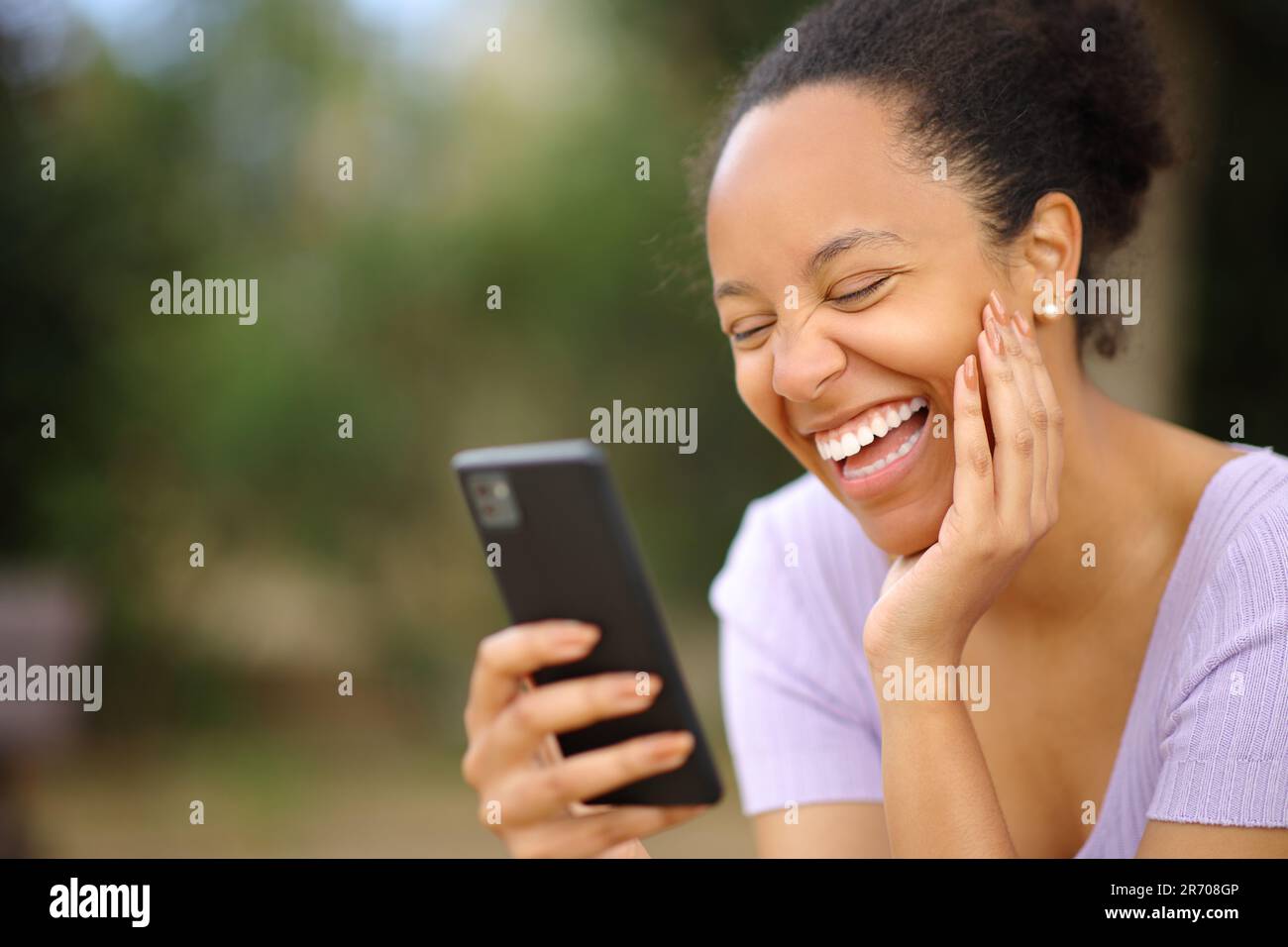 Divertente donna nera che controlla il telefono ridendo forte in un parco Foto Stock
